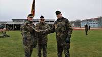 Drei Soldaten in Flecktarn-Uniform stehen auf der Wiese und reichen sich die Hände zu einem Handschlag. 