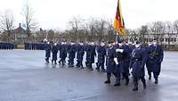 Mehrere Soldaten in blauen Uniformen mit Mänteln marschieren auf einem Platz und tragen dabei die deutsche Flagge.