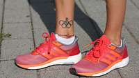 Zwei Beine bekleidet mit orangenen Sportschuhen, am rechten Knöchel ein Tattoo: die olympischen Ringe mittig mit einer Rose.