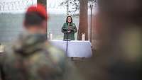 Die Militärpfarrerin predigt unter freiem Himmel vor einem Altar, hinter ihr ein Zaun aus Stacheldraht, im Vordergrund Soldaten