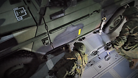 Zwei Soldaten liegen und hocken in einem Flugzeug neben einem grünen Geländewagen.