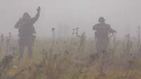 Zwei Soldaten bewegen sich nebeneinander durch dichten Nebel auf einer Wiese.