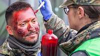 Eine Soldatin malt einem Soldaten Wunden ins Gesicht, damit dieser einen Verwundeten darstellen kann.