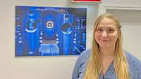 Ein blonde junge Frau lächelt in die Kamera. Sie steht vor einem Bild, welches Lasertechnologie zeigt.