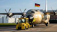 Ein unlackiertes Flugzeug vom Typ C-130J mit Deutschlandflagge wird über das Produktionsgelände gezogen.