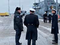 Marinesoldaten in dunkelblauen Uniformmänteln stehen auf einer Hafenpier; im HIntergrund zwei kleine, graue Kriegsschiffe.