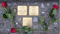 Blick auf drei Bronzeplatten im Straßenpflaster mit Namen von Opfern des Hitlerregimes und Rosen