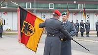 Ein Soldat übergibt eine schwarz-rot-goldene Fahne mit Bundesadler an einen zweiten. Dahinter stehen weitere Soldaten.