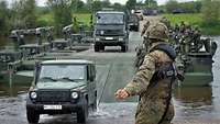 Militärfahrzeuge überqueren eine Militärbrücke über einen Fluss. Ein Soldat weist sie ein.