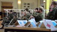 Mehrere Soldaten sitzen in einem Saal und lauschen einem Vortragenden zu. Im Vordergrund stehen auf einem Tisch Taktikkarten.