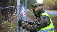 Ein Soldat im Feldanzug und Warnweste befestigt etwas an einem Bauzaun. 