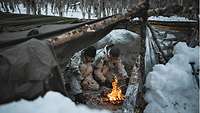 Zwei Soldaten in Schneetarnanzügen sitzen an einem kleinen Feuer hinter einem Schneewall.