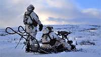 Ein Soldat sitzt in einer Schneelandschaft hinter einem Maschinengewehr. Ein zweiter steht daneben.