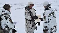 Ein Soldat im Schneetarnanzug überreicht einem zweiten eine Urkunde, ein dritter Soldat schaut zu.