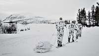 Vier Skiläufer in Schneetarnanzügen ziehen einen Schlitten durch eine tief verschneite Landschaft.