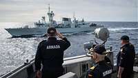 Marinesoldaten in blauer Uniform mit dem Rücken zum Betrachter grüßen militärisch; im Hintergrund ein graues Kriegsschiff in See