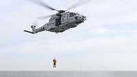 Ein grauer Hubschrauber fliegt durch die Luft und hat eine Person an einem Seil hängen.