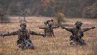 Drei Soldaten auf einer Wiese, hinter ihnen der Ausbilder. Er überprüft die Ausrüstung.