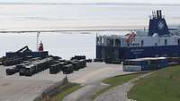 A vehicle ferry docks at a wharf and is loaded with various military vehicles.