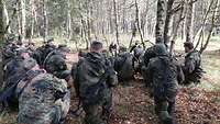 Soldaten in Flecktarn hocken am Boden in einem Wald.