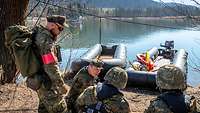 An einem See hocken Soldaten vor zwei Schlauchbooten, daneben ein Ausbilder mit roter Armbinde. 