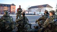 Eine Gruppe Soldaten sitzt in einer Kaserne am Boden. Einer steht vor ihnen und erklärt etwas.