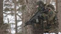 Ein Soldat mit einem Gewehr kniet im Schnee neben einem Baum im Wald.