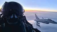 Ein Pilot macht ein Selfie im Cockpit eines Eurofighters während des Flugs. Hinter ihm fliegt ein Eurofighter über den Wolken. 