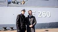 Ein Marineoffizier in dunkelblauer Uniform und ein Zivilist in grauem Mantel stehen vor einer grauen Schiffswand.