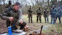 Ein Soldat führt Kameradinnen und Kameraden die Zubereitung eines Gerichtes am Lagerfeuer vor.