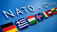 Unter dem Schriftzug NATO – OTAN sind mehrere Flaggen von Mitgliedsstaaten zu sehen.