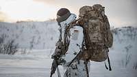 Ein Soldat mit Rucksack und Waffe läuft auf Skiern in einer verschneiten Landschaft.