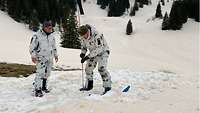 Zwei Soldaten im Schneetarnanzug suchen mit einer Sonde, einem schmalen Stab, im Schnee.