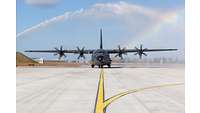 A C-130J taxies on the runway. Water jets can be seen above it. A rainbow is formed by the sun.