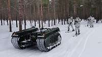 Ein unbemanntes Kettenfahrzeug zieht vier Soldaten in Schneetarn auf Skiern über einen Waldweg.