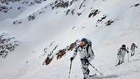 Soldaten im Schneetarnanzug steigen bei Sonne mit Ski einen weiß verschneiten Berg hoch.