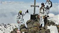Drei Soldaten stehen auf einem felsigen Berggipfel über den Wolken neben dem Gipfelkreuz im Licht.