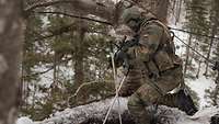 Ein Soldat kniet hinter einem Baum und schießt mit dem Gewehr den Hang runter.
