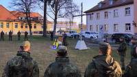 Auf einer Freifläche blicken Soldaten zu einem Pfarrer in Uniform, der vor einem Birkenkreuz steht.