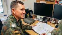 Ein Soldat sitzt an seinem Schreibtisch und blickt zu einem weiteren Soldaten auf 