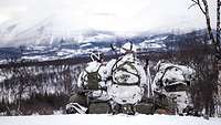 Drei Soldaten in Schneetarnuniform sitzend von hinten. Im Hintergrund eine weitläufige Winterlandschaft mit Bergen am Horizont.