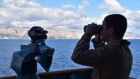 Ein Soldat steht mit einem Fernglas an einer Reling und schaut auf das Meer, im Hintergrund Berge