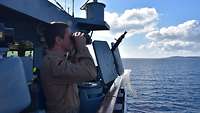 Ein Soldat steht auf dem Deck eines Schiffes und schaut durch ein Fernglas auf das Meer