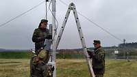 Soldatinnen und Soldaten stehen auf einem Feld um eine Leiter herum, die an einen Mast gelehnt ist.