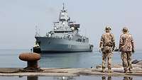 Zwei Personen in Uniform stehen auf einer Pier in einem Hafen und schauen auf ein großes graues Schiff.