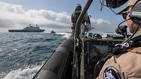 Detailaufnahme vom Fahrstand auf einem Speedboot in See; im Hintergrund ein großes, graues Schiff.