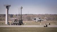 Ein Hubschrauber, knapp über dem Boden schwebend, und ein gepanzertes Fahrzeug mit Antennen sind auf dem Flugplatz in Jagel.