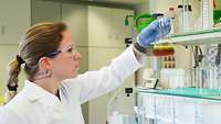 Eine Frau mit Brille und weißem Arbeitskittel untersucht eine Flüssigkeit in einem Labor