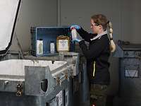 Eine Frau in dunkler Arbeitskleidung mit Zopf und Brille untersucht in einem Raum Gefahrstoffe an einem Schutzcontainer