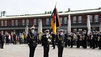 Drei Männer in Uniform stehen auf einem gepflasterten Platz. Der Mann in der Mitte hält eine Fahne.